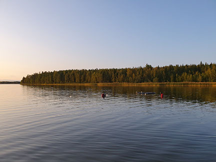 Två personer med dykarkläder står i vattnet i solnedgången, i bakgrunden syns träd.